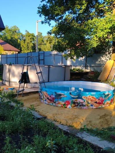 This media file depicts a backyard setting in Kotovsk, Russia, with two above-ground swimming pools. The foreground features a smaller, round pool decorated with vibrant aquatic life, including sea turtles and dolphins. The pool is filled with clear blue water. Adjacent to it is a larger, rectangular pool, currently illuminated by sunlight, with a gray exterior and a ladder with a towel draped over it.
The scene is set on a sunny day, indicated by the bright sunlight and clear blue sky. Lush green trees and foliage surround the pools, suggesting a warm, possibly summer, season. In the background, a light-colored fence runs along the property line, separating the yard from neighboring areas. A house with a dark-tiled roof is partially visible on the left, and a lamppost stands tall in the distance. The ground around the pools is covered in sand and some patches of green grass and flowering plants.
There is no activity of people or animals visible in the media file. The overall atmosphere is serene and suggests a leisure setting, possibly prepared for outdoor recreation. The sunlight casting shadows from the trees and illuminating the pools indicates that the photo was likely taken in the late afternoon or early evening.