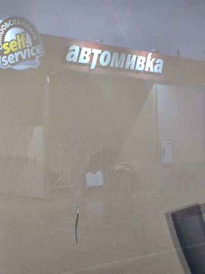 The image captures an outdoor scene of what appears to be a self-service car wash in Kraishte, Bulgaria. The primary subject is a building with prominent signage.
A large, brightly lit sign on the building displays the Cyrillic word "автомивка" (avtomivka), which translates to "car wash." Below this, a circular logo is visible, featuring the English text "Self Service" and the corresponding Bulgarian word "САМООБСЛУЖВАНЕ" (samoobsluzhvane), also meaning "self-service." No people or active events are discernible in the frame, presenting a static view of the facility.
The scene is set under overcast conditions, indicated by the diffused, even lighting and the muted grey-blue sky. There are no strong shadows, suggesting either a heavily clouded day or possibly dusk/dawn. The ground surface appears to be light-colored concrete or asphalt, showing some reflections or texture variations. A dark, rectangular object, possibly a grate or vent, is partially visible in the upper right corner of the image. The photograph exhibits a grainy texture and slight blurriness, contributing to a muted overall color palette. The image is oriented vertically, suggesting it was taken from a tilted perspective.