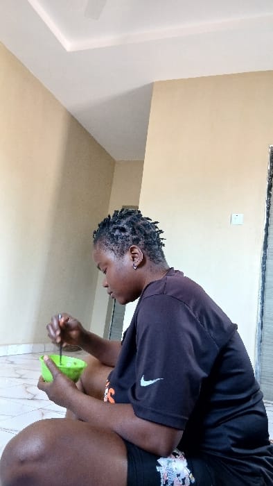 An individual with dark skin and short dreadlocks is observed seated on a tiled floor indoors in Bashar, Nigeria. The person wears a dark short-sleeved t-shirt featuring a white Nike swoosh logo on the left chest and dark shorts with a pattern on the right thigh. A silver-colored hoop earring is visible in the left ear. The individual holds a bright green bowl in their left hand and uses a dark-colored utensil with their right hand to stir or consume the light green substance within the bowl. The room features light beige walls, a white ceiling with a recessed panel design, and a white and grey marbled tiled floor. A white ceiling fan is partially visible in the upper left corner of the ceiling. A white light switch or power outlet and a dark door frame are visible on the right side of the image.