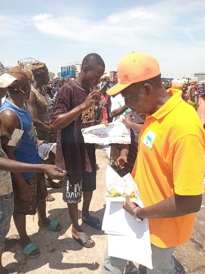 The scene takes place in an outdoor setting in Katabu, Nigeria (latitude: 10.71, longitude: 7.514). A group of people are gathered in a busy area, possibly a market or a public space. A man in a bright orange shirt and hat is distributing flyers to a group of people. He is holding a stack of flyers in his left hand, and he is pointing at one of the flyers with his right hand. He appears to be explaining the flyer to the people he is giving them to, and they are listening attentively. Some of the people are holding the flyers in their hands. There is a person wearing a dark-colored shirt standing in front of the man in the orange shirt, he is also holding a flyer. He is holding a cell phone in his right hand and pointing it at the man in the orange shirt with his left hand. Several other people are present in the background, including some who are looking at the flyers and some who are talking to each other. The environment is dusty and rough, with dirt on the ground.