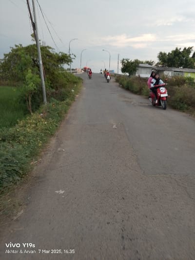 The image shows a paved road with several motorbikes driving on it. The road is straight and slopes gently upwards. There are a few trees and shrubs on both sides of the road. A small group of people can be seen on a motorbike in the distance. The sky is cloudy and overcast. The image appears to be taken from a moving vehicle looking toward the front. There are multiple power lines running along the road, suggesting it is an urban area. The coordinates provided (-6.375, 108.055) place the location in Kecamatan Gabuswetan, Indonesia.