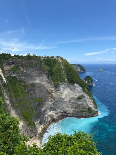 This image showcases a dramatic coastal landscape at Nusa Penida Island in Bunga Mekar, Indonesia. Dominating the scene is a towering, layered cliff face with varying shades of grey and dark rock. The upper portion of the cliff is covered in lush, green vegetation. A narrow path is visible along the cliff edge. Below the cliff, the turquoise ocean meets the rocky coastline. Two smaller, rocky islands with vegetation are situated in the deep blue sea. A boat is travelling near the islands. The sky is a clear blue with a few wispy clouds. Green foliage is prominent in the bottom right of the frame.