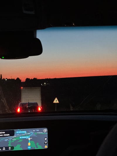 The scene shows a view from inside a car, likely at dusk or night. The sky outside the window displays a gradient of light blue to peach or pink. In the car's rearview mirror, a truck with illuminated red taillights is visible. A road sign indicating an upcoming intersection or merge is present. Inside the car, a navigation system displays a map of the area, showing a route through Ciucur-Mingir, Moldova. The navigation system highlights street names such as "Str. Barbu Lautaru" and "Str. Cetatea Alba", and estimates a remaining distance of "5,2 km" and a travel time of "69 minutes". The current speed is noted as "72 km". The car is either stopped or moving slowly based on the navigation information. The focus is on the road ahead and the navigation system's guidance.