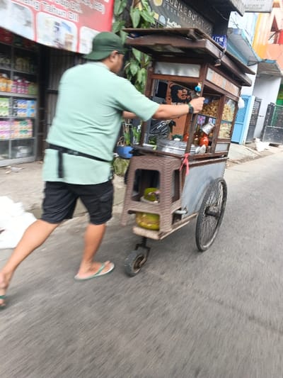 A male individual, seen from behind, pushes a three-wheeled street vendor cart along a paved road in Dayeuhkolot, Indonesia. The man wears a light green short-sleeved shirt, dark shorts, a dark belt, a dark green baseball cap, and light green flip-flops. A watch is visible on his left wrist. The cart, constructed from wood and metal, features an upper display section with glass panels containing various packaged snacks and bottles. A large metal pot is situated on the cart's counter. A brown plastic stool, affixed to the lower front of the cart, holds a green gas cylinder. A red strap is visible near the gas cylinder. The cart utilizes two large bicycle-style wheels on the right side and a smaller castor wheel at the front left. On the left side of the road, a storefront displays packaged goods on shelves beneath a red banner with partial white text. A green plant grows next to the building. On the right side, a building with light blue and orange exterior elements and a metal fence is visible. The scene is illuminated by ambient light, with motion blur indicating the man and cart are actively moving forward.