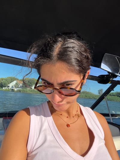 The image displays a female individual situated in a boat on a body of water within the city of Coruche, Portugal. The subject, a woman with dark, wavy hair pulled back, occupies the central foreground, looking directly at the camera. She wears tortoise-shell framed sunglasses positioned low on her nose, enabling her eyes to be seen above the lenses. Her facial expression is direct, with a subtle downturn of the mouth. She is dressed in a light-colored, possibly white or pale pink, ribbed tank top. Around her neck are two gold-toned necklaces; one features small red beads, and the other a single rectangular reddish-brown pendant. The scene is brightly lit by sunlight, highlighting her tanned skin.
In the background, a clear blue sky is visible above the water. The boat's canopy is partially visible at the top, and a portion of the boat's windshield or frame runs across the middle ground. To the subject's left, across the water, a distant shore features several white buildings with reddish roofs and surrounding green foliage, suggesting a residential or village area. To her right, part of the boat's internal structure, including a black pole-mounted mirror, is visible. The water surface exhibits ripples, indicative of a gentle current or boat movement.