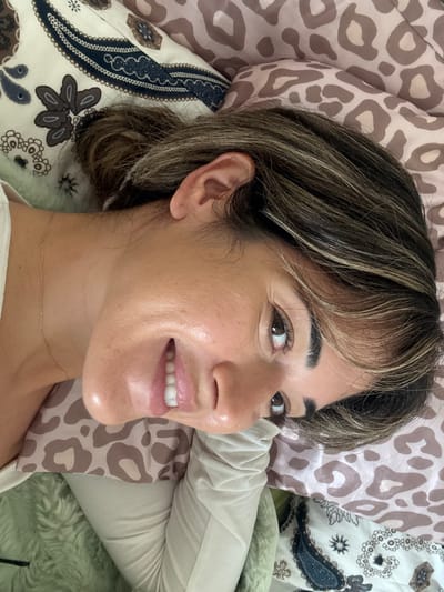 An image taken indoors, likely a bedroom, within the Kalyvia Thorikou Municipal Unit, Greece, depicts one individual. A woman with dark brown hair and blonde highlights is positioned on her left side, her head resting on a pillow. Her face is directed towards the viewer, exhibiting a smile with visible white teeth. Her eyes are open and dark brown. A thin, gold-colored chain necklace is visible around her neck. She appears to be wearing a light-colored, possibly cream, long-sleeved garment. The bedding includes a pillow with a brown leopard print pattern directly under her head and another pillow with a cream background and a dark blue and brown paisley design to her upper left. A light green, textured fabric, possibly a blanket, is partially visible to her lower left. The image is rotated clockwise, placing the woman's head towards the upper right quadrant of the frame.
