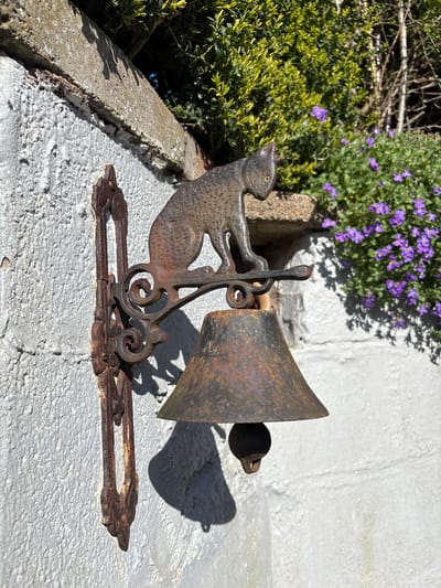 The image shows a rusty, cast iron cat-themed door bell. The cat figure is attached to a decorative bracket holding a weathered, cone-shaped bell with a spherical clapper. The bell assembly is mounted on a whitewashed brick wall, likely a fence or the side of a building. Adjacent to the bell are green bushes and purple flowers. Given the prompt, this scene is presumed to be located in Snailbeach, United Kingdom.