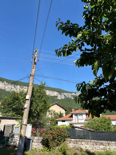 The image shows a view from a residential area in Pavolche, Bulgaria, with a clear blue sky. In the foreground, a weathered concrete utility pole stands prominently, carrying multiple overhead wires. The pole is equipped with various electrical components, including insulators and what appears to be a small transformer or junction box. To the left of the pole, a section of a property is visible, featuring a building with a green gate and a stone wall. Adjacent to this, a dense bush with pink flowers adds a touch of color. Behind the pole and flowers, more houses with red tile roofs and green trees are scattered across the landscape. The background is dominated by a majestic, rugged mountain range covered in lush green vegetation, with a distinct cliff face visible on its upper slopes. A large tree with dark green leaves partially obscures the right side of the image, casting shadows. The scene depicts a typical day in a Balkan village, characterized by its connection to nature and the presence of essential utilities.