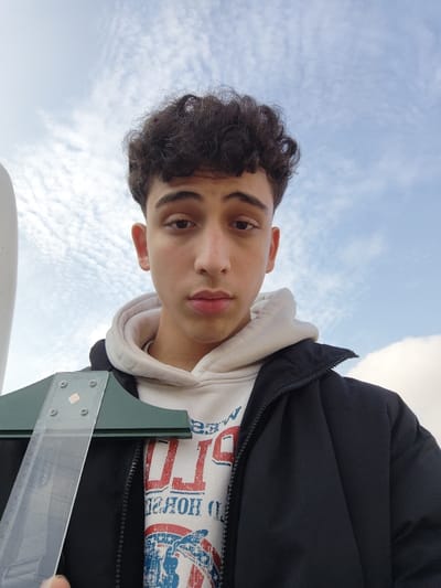 An image taken in Mankat al-Hatab, Syria, depicts a young male, appearing to be a teenager or young adult, positioned centrally and looking directly into the camera. He possesses curly dark hair, light-to-medium skin, and is wearing a light-colored hooded sweatshirt under an unzipped dark puffer jacket. A partial graphic print featuring red and blue elements, possibly including text, is visible on the sweatshirt. The subject holds a green T-square, a drafting tool, in the lower left portion of the frame, with its transparent ruler component and metallic fasteners clearly visible. A portion of a white vertical structure is present on the far left. The background consists of an outdoor sky featuring scattered altocumulus or stratocumulus clouds. The lighting suggests daylight conditions.