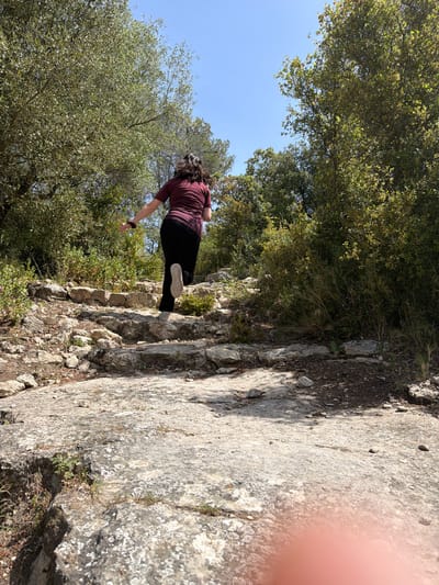 A person is captured from behind, walking or stepping carefully on a rocky, uneven path in Olèrdola, Spain. The individual wears a maroon short-sleeved top, black trousers, and light-colored sneakers. Their dark hair is pulled back. The path is made of large, light-colored rocks, some covered with lichen, and dry earth with sparse vegetation like grass and small bushes.
The scene is an outdoor, natural environment, likely a mountainous or hilly area. Dense green trees and foliage flank both sides of the path, with a clear, bright blue sky visible above and between the tree canopies. The strong sunlight suggests it is midday or early afternoon, casting distinct shadows on the rocky ground. The camera is held at an angle, making the ground appear significantly tilted. A blurry, pinkish area on the far right edge indicates a finger partially obstructing the lens. No text is visible in the image.