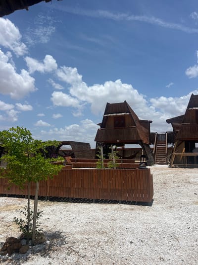 The image shows two wooden structures, both with pitched roofs, located in Cabo de la Vela, Colombia, at coordinates 12.201 latitude, -72.148 longitude. The structures are partially obscured by a wooden fence and a small tree in the foreground. The structures are built in a traditional style with a thatched roof and a wooden frame. The ground is covered in white sand and pebbles. The sky is blue with white clouds. There is a slight overhang of a thatched roof in the upper left corner of the image. The structures appear to be part of a small resort or lodging complex.