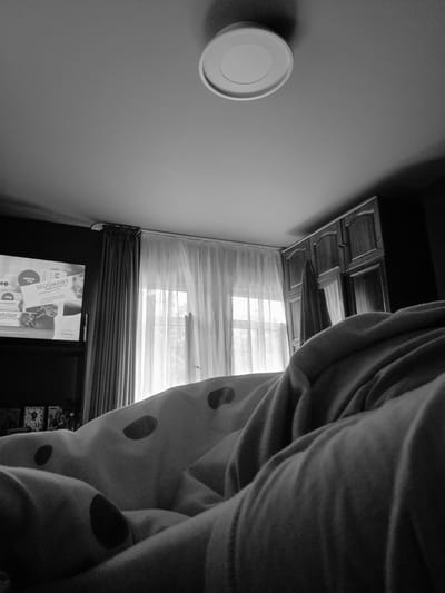 In a dimly lit bedroom, a black and white, low-angle shot focuses on a person lying in bed, under a blanket with a polka-dot pattern. The blanket fills the foreground, obscuring most of the person's body. In the background, a wardrobe with dark wood doors stands against the wall, with a draped towel hanging from one of the doors. To the left of the wardrobe, a window is covered by sheer white curtains, allowing diffused natural light to enter the room. To the left of the window, a dark curtain is drawn, and a television screen displays an advertisement for a medication. The ceiling features a white circular light fixture. The scene is likely a bedroom in a residential building in Iskrets, Bulgaria.