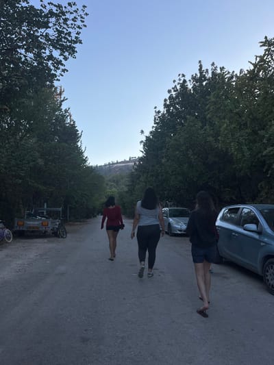 Three individuals walk away from the camera along a paved road in the Merom HaGalil Regional Council, Israel, under a clear, light blue sky during what appears to be late afternoon or early evening. The person on the left wears a dark red long-sleeved top and dark shorts, with bare feet. The middle individual wears a light-colored short-sleeved top and dark leggings with light-colored shoes. The person on the right wears a dark long-sleeved top and dark shorts, with dark flip-flops. All three have dark hair.
The road is bordered on both sides by dense green trees and foliage. On the left side, a white flatbed trailer with a visible Israeli license plate is parked, alongside a purple-framed bicycle and a yellow/black bicycle. On the right side, two passenger cars are parked: a light blue/silver hatchback is closer to the foreground, and a silver sedan is parked further down the road. In the distant background, a tree-covered hill or mountain is visible against the sky.