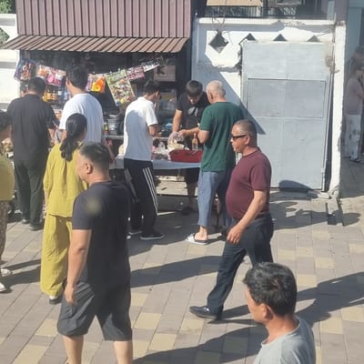 A group of people are gathered around a small outdoor stall selling various items, likely toys and snacks, under a brown awning. The stall is set up in a paved outdoor area with a checkered pattern of light and dark square tiles. Behind the stall is a building with a corrugated metal roof and a light grey metal gate.
In the foreground, from left to right, a person in a yellow outfit stands with their back to the camera. To their right, a man in a black t-shirt and grey shorts is walking away from the viewer. Further right, a man in a maroon t-shirt and dark jeans walks past, looking towards the left. Another man in a grey t-shirt is partially visible in the far right foreground, his head turned away from the camera.
Near the stall, several people are interacting. A man in a green t-shirt and blue pants is handing something from a red container to a man in a black t-shirt. Another man in a white t-shirt stands nearby, looking towards the stall. A woman in a black t-shirt and patterned pants is on the far left, mostly obscured.
The scene appears to be a market or street-side vendor location. The overall impression is a busy, casual street scene. This is a scene consistent with the general street vending environments that can be found in the city of Turkistan, Kazakhstan, particularly in areas with pedestrian traffic and smaller commercial establishments.
