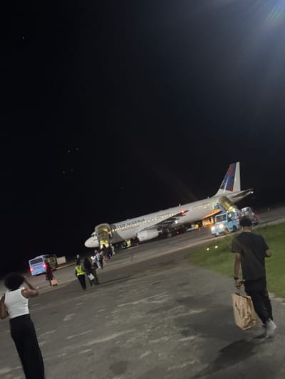 At Omoecheigbo, Nigeria, an airplane with "UNITED NIGERIA" on its side is parked on a tarmac at night. People are near the plane, some standing, and some near vehicles. The environment is dark, with some artificial lighting illuminating the scene.