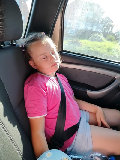 A young girl with blonde hair styled in pigtails with colorful clips is sleeping in the back seat of a car. She is wearing a bright pink t-shirt with white trim and light blue shorts. A black seatbelt is fastened across her chest. The girl's eyes are closed, and her head is resting against the car's headrest. Sunlight streams in from the window, illuminating her face and casting shadows on the dark gray car seat. Outside the window, green foliage and a building are visible through the slightly dirty glass, suggesting a daytime scene in a rural or suburban area. A water bottle lies on the seat in front of her. The image appears to capture a moment of rest during a car journey.