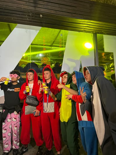 The image captures a group of six young men posing for a photo in an indoor setting. The individuals are wearing various costumes, including a shark, Captain America, a dinosaur, two red jumpsuit-like costumes, and one wearing Hello Kitty pajama bottoms and a black t-shirt emblazoned with "Pornhub". They appear to be at a party or gathering, as indicated by the festive attire and the presence of cylindrical containers, possibly drinks or party favors. The background features a mix of wood slats, mirrors, and reflective surfaces with geometric designs. The interior lighting has a yellowish hue. It is not possible to determine the location in the city of Grau I Platja, Spain, based on visual information.