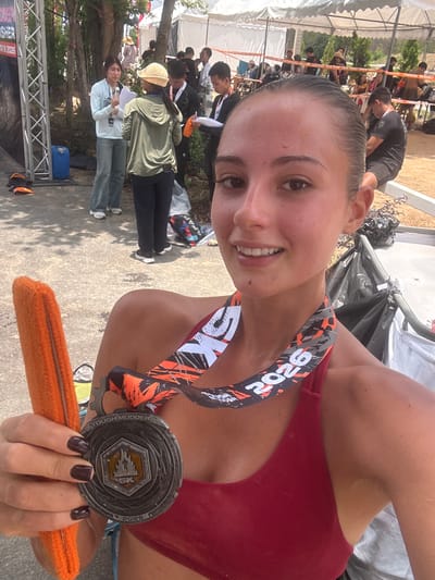 A selfie taken in บ้านสำนักตะแบก, Thailand, depicts a woman with light skin, dark hair pulled back, and wearing a red sports bra. She holds a "Tough Mudder 5K 2026" medal in her left hand, along with an orange headband. The medal hangs around her neck with a black, white, and orange patterned ribbon. The woman is smiling and looking at the camera.
The background shows an outdoor setting with people and structures. There are figures wearing various clothing, and a tent structure with an orange barrier is visible. Some trees and foliage can also be seen. The ground appears to be paved with concrete or asphalt.