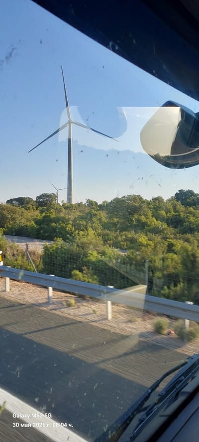 A partially obstructed view shows wind turbines, foliage, a guardrail, and a road. The view is likely from inside a vehicle, evidenced by reflections on the windshield and a side mirror. The time stamp on the image indicates May 30, 2026. Based on the presence of wind turbines, this scene is located in Tébar, Spain.