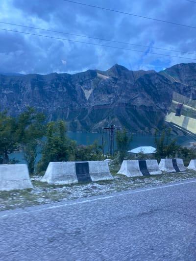 The image depicts a mountainous landscape with a body of water. In the foreground, a paved road is visible, bordered by concrete barriers with black and white markings. Beyond the barriers, there are scattered rocks and vegetation, including several trees. The body of water, appearing to be a lake or reservoir, is turquoise in color and is situated in a valley. Towering mountains with rugged slopes rise behind the water. The sky is cloudy and a bluish-gray color. Power lines are strung across the upper portion of the image. Reflections are visible on the surface of the water and on what appears to be a window, suggesting the photo was taken from a moving vehicle. The scene is likely located in or near the village of Irganai, Russia, given its mountainous terrain and the presence of a reservoir, which is consistent with the region's geography. There are no people visible in the image.
