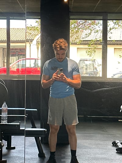 A man stands facing his reflection in a mirror in a room that seems to be a gym. He is fair-skinned with light brown hair, wearing a blue t-shirt and grey shorts. He is holding a smartphone in his hands, pointed toward the mirror. The reflection behind him shows a building across the street with cars parked in front. One car's license plate appears to read "WÜ CI 5052". Based on the license plate, the city is Jestetten, Germany. In the gym, there is workout equipment visible near the man. A water bottle is seen near the bottom of the image.