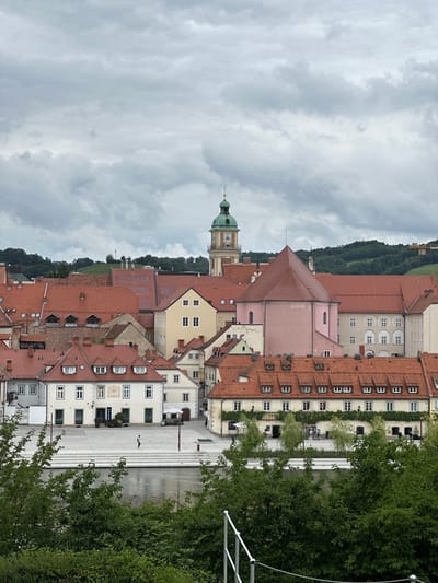 This is a photograph of the Lent district in Maribor, Slovenia, a historic riverfront area. The scene depicts a collection of buildings with red tile roofs, typical of European architecture, lining a wide plaza. In the background, a prominent clock tower with a green dome stands out against a cloudy sky. The plaza in the foreground features white paving stones and a series of stepped platforms leading down towards a body of water, likely the Drava River, which occupies the lower portion of the frame. Lush green trees and foliage in the foreground partially obscure the view of the plaza and river. A single person is visible walking across the plaza. The architecture suggests a mix of historical and more modern structures. The overall impression is a charming, somewhat overcast, urban landscape.