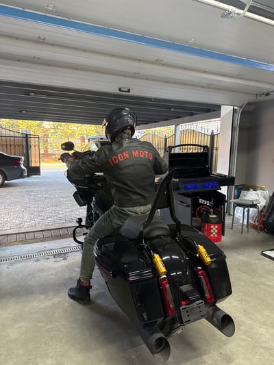 A person wearing a black motorcycle helmet and a black jacket with the words "ICON MOTO" in red on the back is sitting on a black motorcycle in what appears to be a garage in Дубровка, Russia. The garage has a gray concrete floor and a white metal garage door with vertical panels. Outside the garage, there is a brick driveway and a view of trees and a metal fence. The motorcycle has black saddlebags and exhaust pipes, with gold-colored shock absorbers. A car is partially visible outside the garage. There is a large black grilling station with blue lights behind the motorcycle.