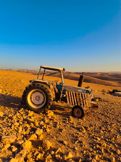 An old, blue and white tractor sits rusting in a barren, rocky landscape. The tractor is the primary subject of the image, with its large rear tire and worn metal body clearly visible. Behind the tractor, rolling, arid hills stretch into the distance, with a small town or settlement visible on the horizon, situated in the Palestinian Territories of Keidar. The sky above is a clear, vibrant blue, indicating a sunny day. The ground is covered in loose stones and sparse vegetation, characteristic of a desert or semi-arid environment. The lighting suggests the photo was taken during either sunrise or sunset, casting long shadows and bathing the scene in a warm, golden light. There are no people visible in the image, and the tractor appears to be abandoned. The overall impression is one of desolation and the passage of time.