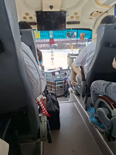 The image shows the interior of a vehicle in บ้านหนองน้ำเต้าลอย, Thailand. Passengers are seated in gray seats. A television screen is visible, and there is luggage in the aisle. One passenger wears blue shoes. A table contains a water bottle and an object with red and white stripes.