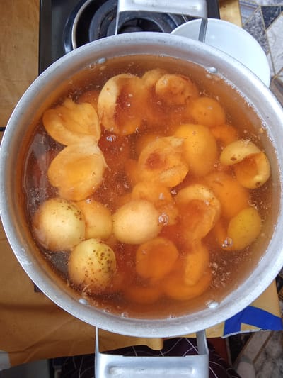 A top-down, close-up shot shows a pot of cooked quince fruit on a stovetop in the city of Жошук, Kyrgyzstan. The quince, peeled and halved, are submerged in a rich, amber-colored syrup, with some pieces showing the characteristic dark specks of the fruit's core. Bubbles rise to the surface, indicating the liquid is still warm. The pot, made of aluminum, is slightly worn and sits on a black gas stove burner. The background is blurred, but hints of a textured countertop and a white bowl are visible. The scene suggests domestic food preparation, likely for preserving or serving the cooked quince.