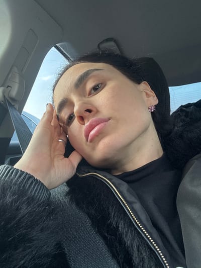 A woman in a car poses for a selfie. She wears a black jacket with fur trim and a black top. Her hair is dark and pulled back. She has pink stud earrings and a silver ring on her left hand. Her hand is near her face, and she is sticking out her tongue slightly. The car's interior is visible, including a seat belt. Bright sunlight filters through the window.