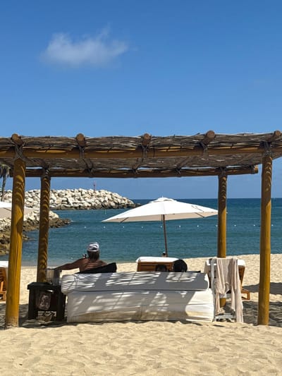 The image displays a sunny beach scene at The Beach, Mexico. In the foreground, a large, rectangular wooden cabana with a woven, thatched roof stands over a white, cushioned daybed. A dark-skinned individual with long hair, wearing a white and dark-striped head covering, is seated on the left side of the daybed, facing the ocean with their bare back visible. To their immediate left, a small wooden side table holds a silver ice bucket containing a lime. To the right of the daybed, a light-colored, striped garment hangs draped over a wooden beach lounge chair. The sandy ground under the cabana exhibits distinct shadow patterns from the roof structure. Beyond the cabana, the calm blue ocean stretches, marked by a line of white buoys. A prominent rocky breakwater extends into the water from the left, featuring a red navigational marker at its far end. Two white beach umbrellas are visible in the midground, with one fully open, shading an unoccupied wooden lounge chair. The background consists of a clear blue sky with a single small white cloud.