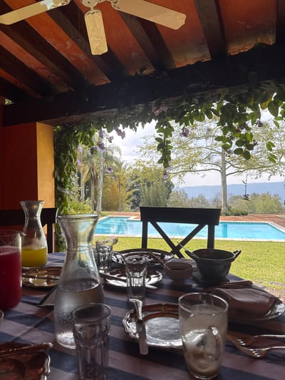 The image captures a breakfast setting on a patio overlooking a swimming pool, possibly in San Pablo Coapan, Mexico. The table is covered with a blue and white striped tablecloth and set with silver serving ware, carafes of juice (one red, one yellow), and filled glasses. A wrought-iron chair is positioned at the table's edge. Beyond the table, a lush garden extends to a bright blue swimming pool. In the background, trees and mountains are visible under a partly cloudy sky. The patio is adorned with flowering vines. A ceiling fan is visible in the upper left corner.