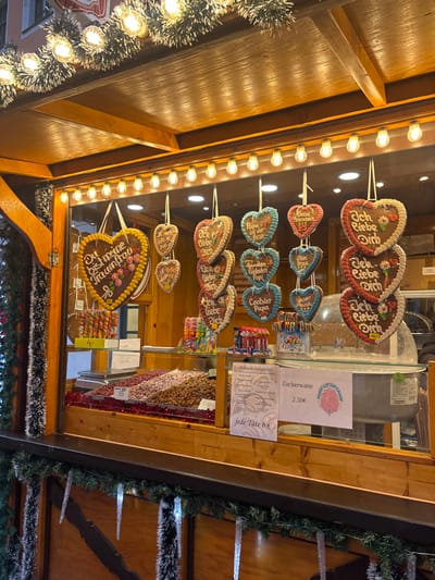 This image captures a festive wooden market stall, likely part of a Christmas market, situated in Meissen, Germany. The stall's exterior is decorated with green tinsel intertwined with warm white string lights along the roof's edge, and illuminated icicle ornaments hang from the lower counter. Inside the brightly lit display window, numerous heart-shaped gingerbread cookies (Lebkuchenherzen) are suspended. These gingerbread hearts feature various German phrases, including "Du bist meine Traumfrau" (You are my dream woman), "Ich liebe Dich" (I love you), "Papa ist der Beste" (Dad is the best), "Kleine Prinzessin" (Little Princess), "Unser Prinz" (Our Prince), "Frechdachs Papa" (Cheeky Dad), and "Coolster Papa" (Coolest Dad). One "Ich liebe Dich" heart also displays "ABV."
Below the hanging cookies, the counter presents several bins filled with various types of nuts or candied nuts, set on a bed of red tinsel. Colorful spiraled lollipops are visible on the left side of the counter, along with smaller packaged candies. White signs are displayed, one advertising "Zuckerwatte" (Cotton Candy) for "2,50€" with a graphic, and another indicating "jede Tüte 6€" (each bag 6€) for other items. A commercial scale is positioned on the left side of the counter, and a cotton candy machine is partially visible on the right. No individuals are present within the frame.