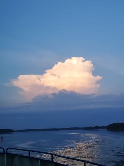 The image captures a serene scene of a body of water, likely a river or a bay, with a distant tree-lined shore. A large, luminous cumulus cloud dominates the upper half of the frame, tinged with warm orange and pink hues, suggesting it is either sunrise or sunset. Below the prominent cloud, darker, lower-lying clouds create a layered effect against the deep blue sky.
The water's surface is calm, with gentle ripples reflecting the sky's colors and the fading light. A faint wake trails behind a vessel from which the photo appears to be taken, indicated by the presence of a railing in the foreground. A solitary red buoy is visible on the left side of the water, marking a channel or hazard.
The overall mood is peaceful and tranquil, evoking a sense of calm and natural beauty. The time of day is clearly either early morning or late evening, with the low-angled light illuminating the clouds. The location is identified as Ul'yanovo, Russia, which aligns with the natural landscape depicted. No text is visible in the image.