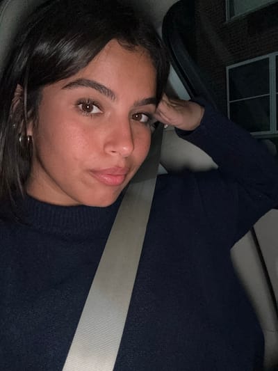 The image displays a female individual with dark brown hair and eyes, seated inside a vehicle during nighttime. She is wearing a navy blue textured sweater and a light-colored seatbelt crosses diagonally from her left hip to her right shoulder. Her left hand is positioned near her head, out of full view. In the background, partially visible through the vehicle's window, is a section of a brick building featuring a white-framed window, illuminated by ambient light. The scene's specific location in Lawrence Township, United States, cannot be confirmed from visual information within the image.
