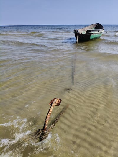 A small, green and black fishing boat is anchored in shallow, clear water near the shore of Lapmežciems, Latvia. The boat is tethered by a thick rope to a rusty anchor lying on the sandy seabed. The anchor is partially submerged in the water, with waves lapping around it, creating small white foam. The seabed is visible through the clear water, showing ripples and patterns in the sand. The sky is a clear blue with a few distant clouds. The overall scene depicts a calm and serene coastal environment.