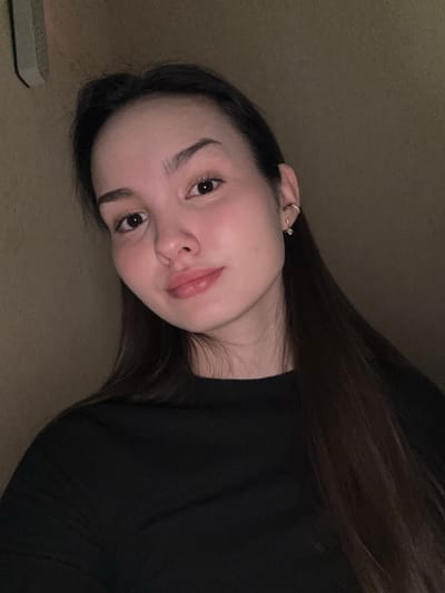 A head-and-shoulders portrait of a young adult female is depicted in an interior setting within Bayronovka, Russia. The subject has long, dark brown hair parted centrally, fair skin, dark brown eyes, and a subtle smile. She is wearing a black, long-sleeved top with a crew neck. On her visible right ear, she has a small gold hoop earring in the upper cartilage and a stud earring with a clear, sparkling stone in the lobe. Her gaze is directed towards the camera. The background consists of a plain, light brown wall. In the upper left quadrant, a portion of an off-white, rectangular object is affixed to the wall. The scene is illuminated by dim, warm lighting, primarily highlighting the subject's face.