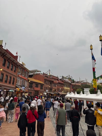 The image shows a crowded public space in Gokarneshwar, Nepal. The scene is oriented with the sky at the top and the ground angled toward the bottom. On the right, people line up along a white structure or platform. Some individuals are prostrating themselves, while others stand or walk. The crowd extends into the lower portion of the frame, filling the walkway in front of brick buildings. The buildings have multiple stories, windows, and some signage in Nepali and English. Above, prayer flags are strung between poles, and a few birds fly in the sky. The sky is overcast and grey.