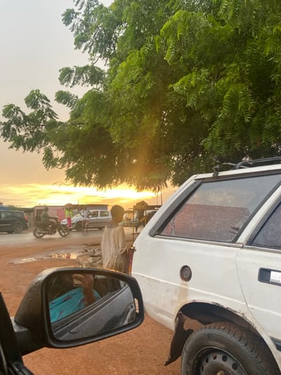 The image captures a vibrant street scene, appearing to be taken from the perspective of someone inside a vehicle, looking out. The overall composition suggests either sunrise or sunset, with warm, golden light illuminating the horizon. The image is oriented vertically, but for description purposes, it will be described as if it were rotated correctly to a horizontal landscape view.

In the immediate foreground, to the right, a black side mirror of a car is visible. The mirror's surface is speckled with water droplets, suggesting recent rain or dew. It reflects a partial view of a person's arm and torso, dressed in a blue or teal patterned garment, and possibly the interior of the vehicle from which the photo is taken.

To the left, a large, lush green tree dominates the frame, its dense foliage creating a canopy. The sun's warm rays break through the leaves, casting a soft glow. The leaves appear slender and numerous, characteristic of certain tropical or subtropical trees.

The midground reveals a dusty, unpaved or poorly maintained road stretching into the distance. A white, possibly older model, van or minivan is prominently positioned, partially obscuring the view of the street beyond. This vehicle shows signs of wear, with visible dirt, possibly rust, and a missing or open fuel cap cover on its side. Its windows appear dusty or wet.

Standing near the white van is a person, seen from the back, wearing light-colored, possibly beige or white, traditional attire. Further down the road, several other vehicles and people can be discerned, including what appears to be another white van or bus, and individuals on bicycles and motorcycles, suggesting active street life. The golden light from the low sun casts long shadows and highlights the dust in the air.

The background is dominated by the sky, displaying a clear gradient of colors from a bright, intense orange and yellow at the horizon where the sun is setting or rising, fading into a lighter, hazier sky above. The overall setting appears to be a rural or semi-urban area, possibly in a warm climate. No specific text is visible in the image.