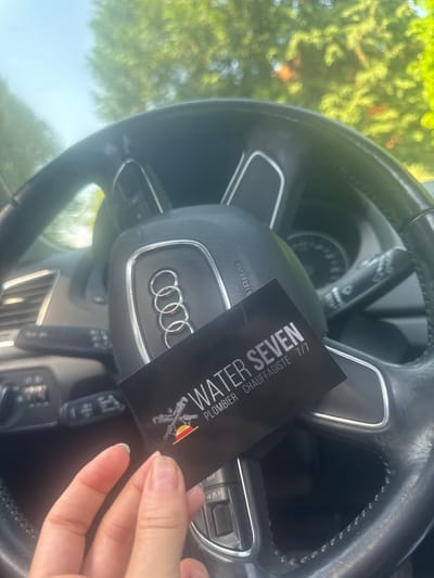 A hand holds a business card in front of an Audi steering wheel. The card reads "WATER SEVEN, PLOMBIER CHAUFFAGISTE 7/7" with a logo of plumbing tools. The environment is the interior of a car with a view of trees and daylight through the windshield. The city and country are not discernible from the image.