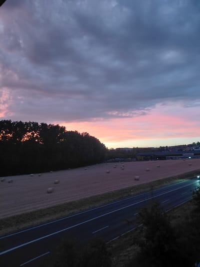 This is an image of a rural landscape at dusk. The sky is a gradient of colors, with dark gray clouds overhead transitioning to shades of pink and orange near the horizon. In the foreground, a mown field is scattered with round bales of hay. A dark road with white lane markings runs diagonally across the lower right portion of the image. Along the left side of the field, a dense line of trees silhouetted against the sky separates the field from a distant town. The town appears to have low-rise buildings, including one with a prominent sign that reads "Boissec". The scene is serene and peaceful, indicative of a rural setting.