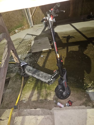 The image captures an outdoor scene, likely a backyard or alleyway, with an electric scooter prominently featured in the center. The scooter is a Segway model, dark gray with red accents, and appears to be in an upright position, possibly leaning against something out of frame or on its kickstand. A yellow hose runs along the ground near the scooter, and a glass is visible on the ground. To the right of the scooter's front wheel, two discarded beverage containers, a glass bottle and a crushed aluminum can, are present. The ground surface is a mix of concrete and dirt, with scattered debris and some mossy patches. In the background, a wooden structure with a thatched roof is visible, and beyond that, dry grass. The overall impression is of an unkempt, possibly urban or semi-rural, outdoor space in Zlataritsa, Bulgaria, with the scooter and debris suggesting recent activity.