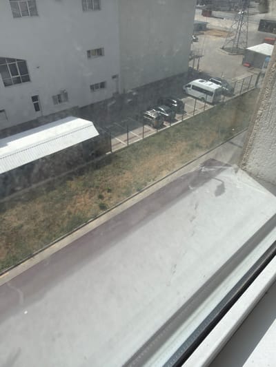 The image provides an aerial view of a parking lot outside a modern, multi-story white building. Several vehicles are parked in designated spots, including a white van, a dark-colored SUV, and a silver hatchback. A chain-link fence separates the parking area from a strip of dry, grassy land. In the background, industrial structures and a tall transmission tower are visible, suggesting an industrial or commercial zone. The scene is captured through a window, with the foreground showing the window sill and pane, which has some smudges and streaks. The overall environment appears to be sunny, with clear daylight.