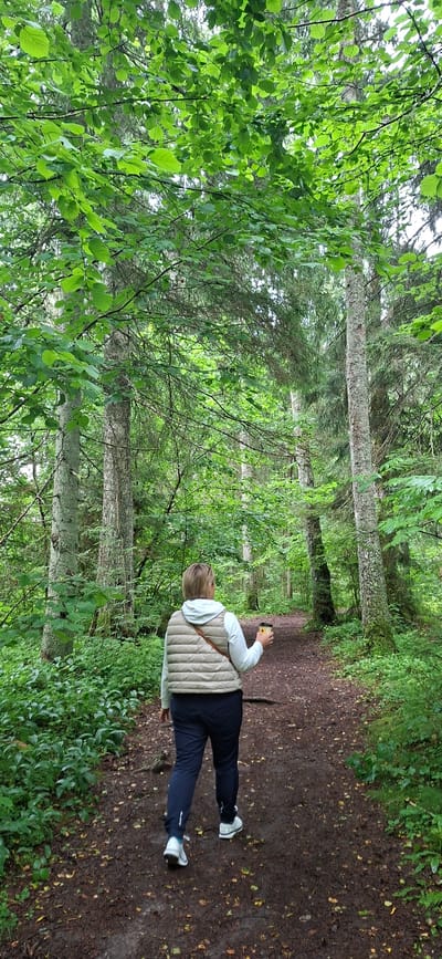 A person walks along a dirt path in a dense forest. The path is lined with trees and green foliage. The person is wearing a light-colored vest, dark pants, and athletic shoes, and is holding a cup in their right hand. The forest appears to be in Saula, Estonia. The scene is peaceful and natural, with the light filtering through the leaves of the trees.