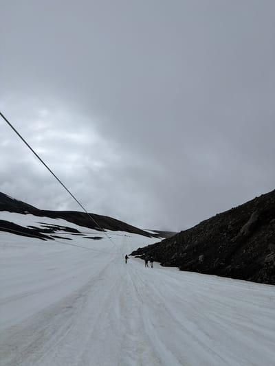 The image shows a cable car in a snowy, mountainous landscape. Several people are visible, appearing to be either riding the cable car or walking on the snow. The environment is barren with dark, rocky slopes and patches of snow. The sky is overcast and gray. The image appears to be taken from a low angle, looking up towards the cable car and the mountain. There is no visible information within the image to suggest this location is in Petropavlovsk-Kamchatskiy, Russia.