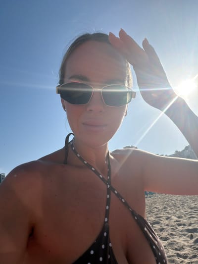 In an outdoor, mid-shot photograph taken on a sunny day at Urbanització Cales I Talaies, Spain, a young woman with blonde hair is pictured. She is wearing sunglasses and a dark brown polka dot bikini. Her right hand is raised, shielding her eyes from the sun, which is a prominent lens flare in the upper right quadrant of the image. The background consists of a sandy beach with some beach chairs and a cliff face in the distance. The woman is looking towards the camera, and her body is angled slightly to the left.