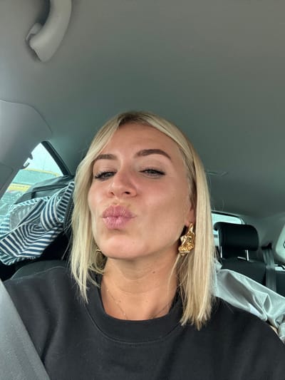 The image displays a close-up selfie of a single individual, likely female, with blonde, shoulder-length hair, seated in the interior of a vehicle. The person has light skin and is looking directly into the camera with lips pursed in a "kissing" gesture. They wear a dark, possibly black, long-sleeved top, a thin gold-colored necklace, and a prominent gold-toned, abstractly sculpted earring on their right ear. A car's grey headliner is visible above, and a dark seatbelt crosses diagonally over their left shoulder and chest. In the rear passenger compartment, a striped fabric item, possibly a bag or clothing, is visible on the left, next to a lighter-colored garment. A blurred view through the back window suggests an overcast sky or an indistinct external environment. The specific location within the city of Brignoles, France, cannot be determined from the visual information provided in the image.