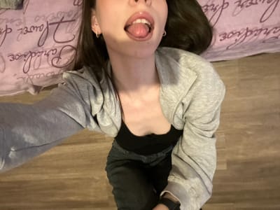 An overhead photograph, taken in Волховец, Russia, depicts a person lying or leaning back on a floor, looking upwards towards the camera. The subject is partially visible, primarily from the chest up, with their head slightly tilted back. Their mouth is open, and the tongue is extended outwards. They have dark hair, and both ears are adorned with small hoop earrings. The individual is wearing a light grey hooded zipper jacket over a black top and dark-colored pants. A dark smartwatch is visible on their left wrist. The immediate background behind the subject's head is a purple textile surface featuring white script text, including the word "Flower" and parts of "Paradise." The area around the subject's body is a light brown wooden floor.