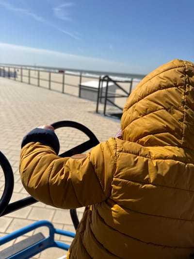 An overhead, slightly rotated photograph depicts a person, likely a child, from behind, wearing a vibrant mustard yellow quilted jacket with a dark blue cuff on the right sleeve. A circular, embossed patch adorns the upper right sleeve. The person's right hand grasps a black, circular handle, possibly part of a play steering wheel or a piece of playground equipment.
The scene is set outdoors under clear, bright daylight, with strong sunlight creating defined shadows on the jacket's folds, suggesting midday or early afternoon. In the background, a paved surface with a herringbone pattern stretches towards the horizon. Beyond this, a light-colored structure, possibly a building or wall, with horizontal metal railings runs across the midground. The sky above is a clear, light blue, subtly fading to a paler hue at the distant horizon, hinting at an expansive, possibly coastal, environment. A very faint crescent moon is just discernible against the blue sky in the upper left quadrant.
Part of a blue, tubular structure, characteristic of playground equipment, is visible in the lower right, reinforcing the context of outdoor play. The overall impression is one of leisure and activity on a sunny day in Middelkerke, Belgium. No legible text is visible in the image.
